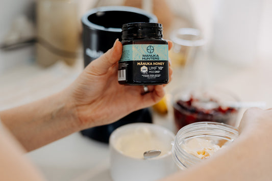 woman's hand holding a manuka hunters jar of honey in the foreground. Healthy breakfast food options in the background. 